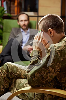 Young distressed woman in military uniform talking to psychologist during therapy session, indoors. Help, support, ptsd