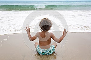 Young diverse boy sitting on the beach doing yoga and meditation.