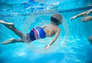 Young diverse boy swimming underwater in a swimming pool. Learning to swim with the help of his parent.