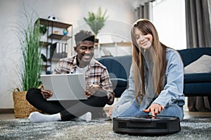 Young diverse couple using robot vacuum cleaner at home