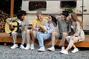 Young diverse friends with pet dog and guitar playing music, having fun near RV on autumn weekend, camping together