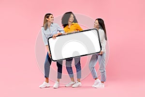 Young diverse women holding giant smartphone with mockup for ad, demonstrating space for your design, full length