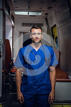 Young doctor in a blue uniform stands and looks at the camera in front of an ambulance car
