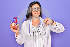 Young doctor cardiology specialist woman holding medical heart over pruple background with angry face, negative sign showing