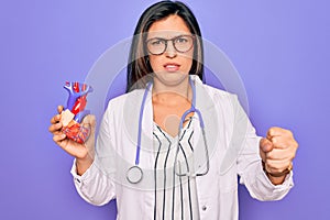 Young doctor cardiology specialist woman holding medical heart over pruple background annoyed and frustrated shouting with anger,