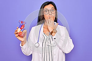 Young doctor cardiology specialist woman holding medical heart over pruple background cover mouth with hand shocked with shame for