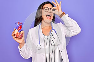 Young doctor cardiology specialist woman holding medical heart over pruple background with happy face smiling doing ok sign with