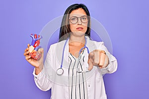 Young doctor cardiology specialist woman holding medical heart over pruple background pointing with finger to the camera and to