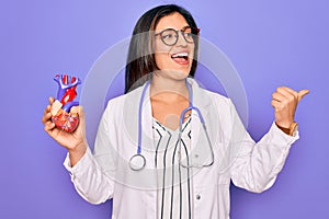 Young doctor cardiology specialist woman holding medical heart over pruple background pointing and showing with thumb up to the