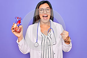 Young doctor cardiology specialist woman holding medical heart over pruple background screaming proud and celebrating victory and