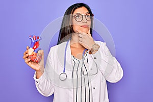 Young doctor cardiology specialist woman holding medical heart over pruple background serious face thinking about question, very