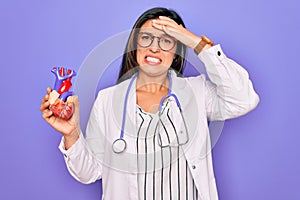 Young doctor cardiology specialist woman holding medical heart over pruple background stressed with hand on head, shocked with