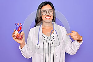 Young doctor cardiology specialist woman holding medical heart over pruple background with surprise face pointing finger to