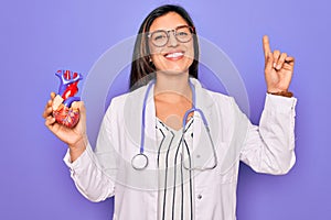 Young doctor cardiology specialist woman holding medical heart over pruple background surprised with an idea or question pointing