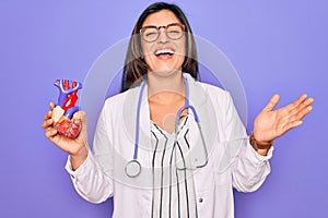 Young doctor cardiology specialist woman holding medical heart over pruple background very happy and excited, winner expression