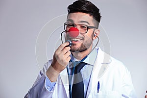 Young doctor with clown red nose listens to himself