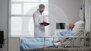 Young doctor in glasses talking to senior man resting in hospital bed. Male RN assisting elderly male patient lying down