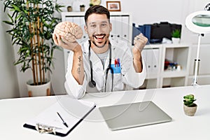 Young doctor holding brain at medical clinic screaming proud, celebrating victory and success very excited with raised arms