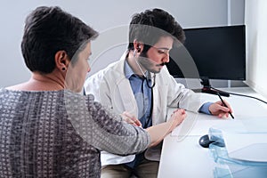 Young doctor listening to mature woman patient heart with a stethoscope and writing down