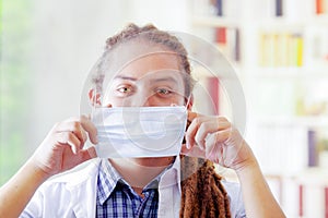 Young doctor with long dread locks posing for camera, adjusting facial mask covering mouth, clinic in background