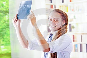 Young doctor with long dread locks posing for camera, holding up x ray image staring at it, clinic in background