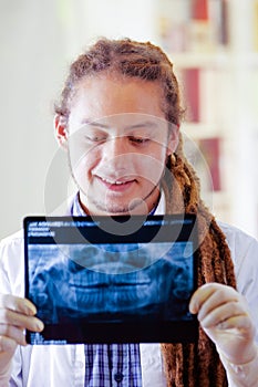 Young doctor with long dread locks posing for camera, holding up x ray image staring at it, clinic in background