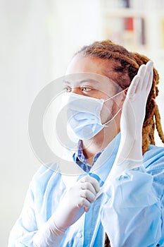 Young doctor with long dread locks posing for camera while putting on rubber gloves, wearing facial mask covering mouth