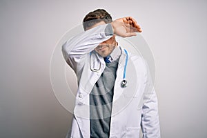 Young doctor man with blue eyes wearing medical coat and stethoscope over isolated background covering eyes with arm, looking
