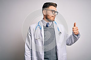 Young doctor man with blue eyes wearing medical coat and stethoscope over isolated background Looking proud, smiling doing thumbs