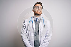 Young doctor man with blue eyes wearing medical coat and stethoscope over isolated background smiling looking to the side and