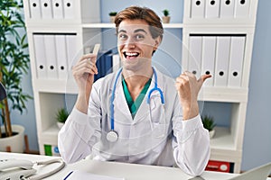 Young doctor man holding band aid pointing thumb up to the side smiling happy with open mouth