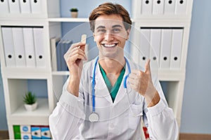 Young doctor man holding band aid smiling happy and positive, thumb up doing excellent and approval sign