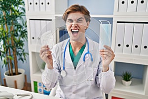 Young doctor man holding two safety mask angry and mad screaming frustrated and furious, shouting with anger