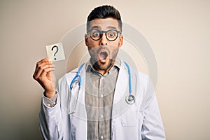 Young doctor man wearing stethoscope holding question mark reminder over isolated background scared in shock with a surprise face,