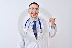 Young doctor man wearing stethoscope over isolated background smiling with happy face looking and pointing to the side with thumb