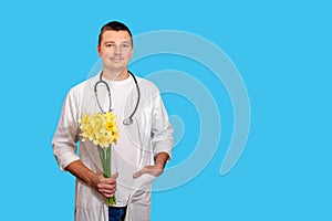 A young doctor men holds a bouquet of yellow narcissists and looks at the camera and stands on a blue background. Concept is