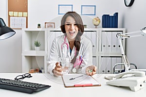 Young doctor woman holding glass of water and prescription pills smiling and laughing hard out loud because funny crazy joke