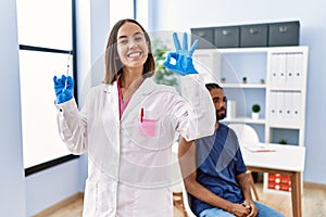 Young doctor woman holding vaccine showing syringe doing ok sign with fingers, smiling friendly gesturing excellent symbol