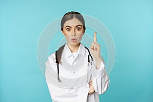 Young doctor, woman physician in white coat raising finger, pointing up, suggesting smth, has solution, revelation