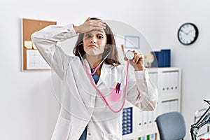 Young doctor woman wearing doctor coat holding stethoscope stressed and frustrated with hand on head, surprised and angry face