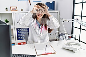 Young doctor woman wearing doctor uniform and stethoscope at the clinic doing ok gesture like binoculars sticking tongue out, eyes