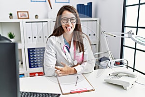 Young doctor woman wearing doctor uniform and stethoscope at the clinic sticking tongue out happy with funny expression