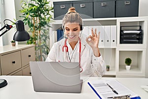 Young doctor woman wearing doctor uniform working using computer laptop doing ok sign with fingers, smiling friendly gesturing