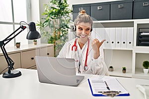 Young doctor woman wearing doctor uniform working using computer laptop pointing thumb up to the side smiling happy with open