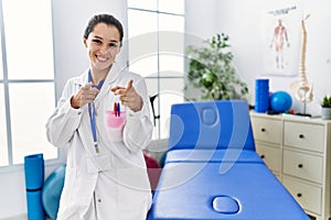 Young doctor woman working at pain recovery clinic pointing fingers to camera with happy and funny face