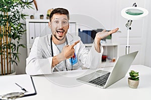 Young doctor working at the clinic using computer laptop amazed and smiling to the camera while presenting with hand and pointing