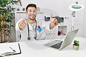 Young doctor working at the clinic using computer laptop approving doing positive gesture with hand, thumbs up smiling and happy
