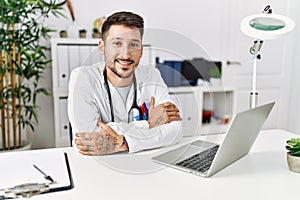 Young doctor working at the clinic using computer laptop happy face smiling with crossed arms looking at the camera