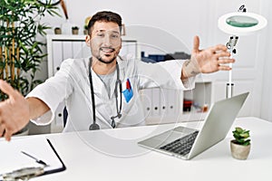 Young doctor working at the clinic using computer laptop looking at the camera smiling with open arms for hug