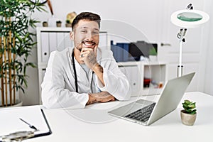 Young doctor working at the clinic using computer laptop looking confident at the camera smiling with crossed arms and hand raised